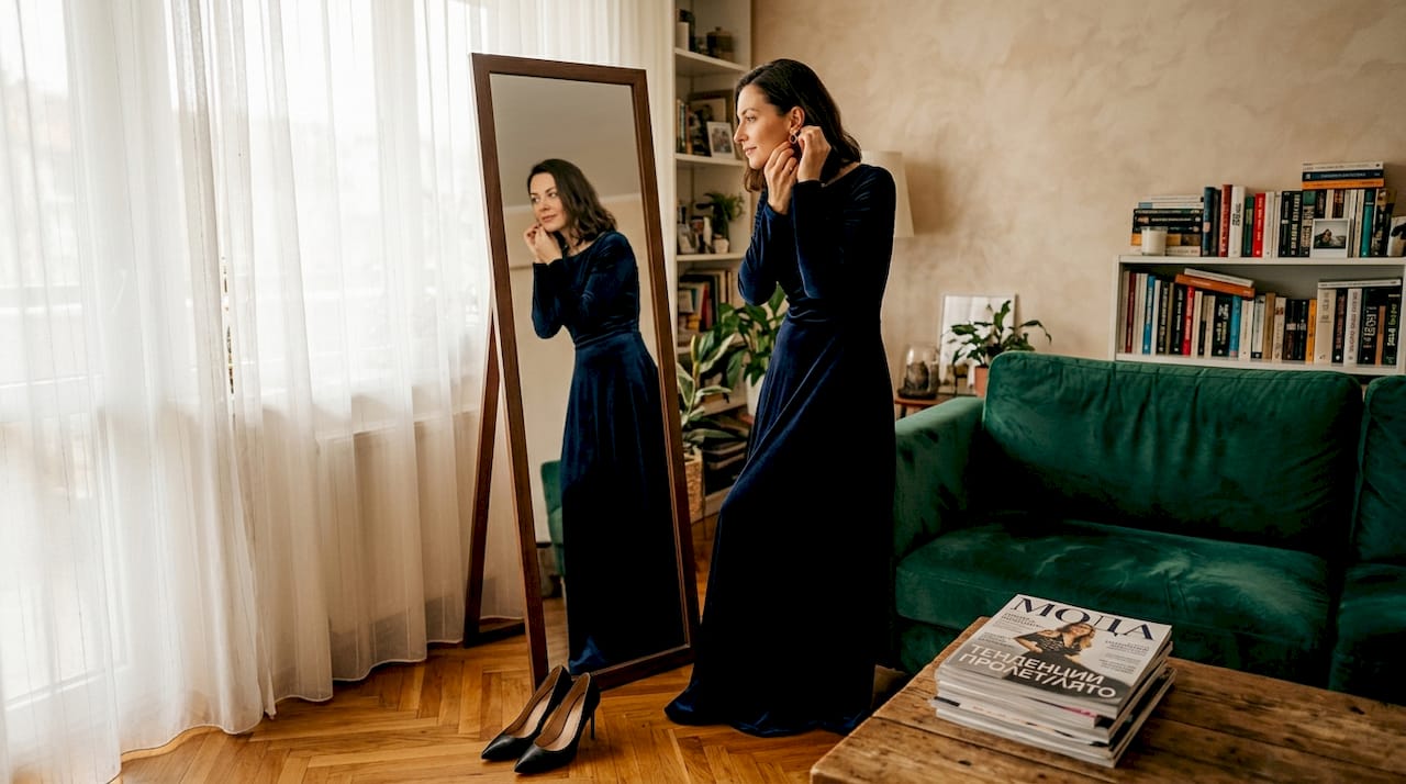 Жена се приготвя за вечерно събитие, обличайки елегантна рокля пред огледалото.